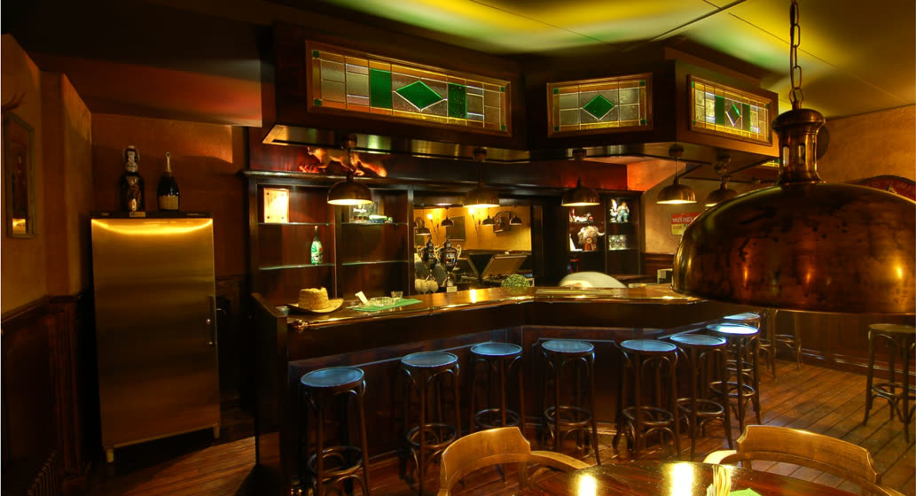 Warme bar met donker hout, glas-in-lood boven de toog en koperen hanglampen